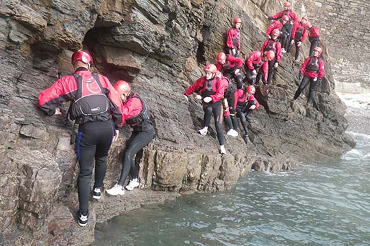 Students in wetsuits and safety gear go coasteering along the shoreline
