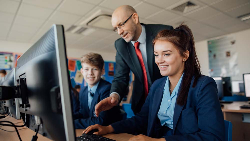 A mature secondary school teacher talks to pupils who are working on computers